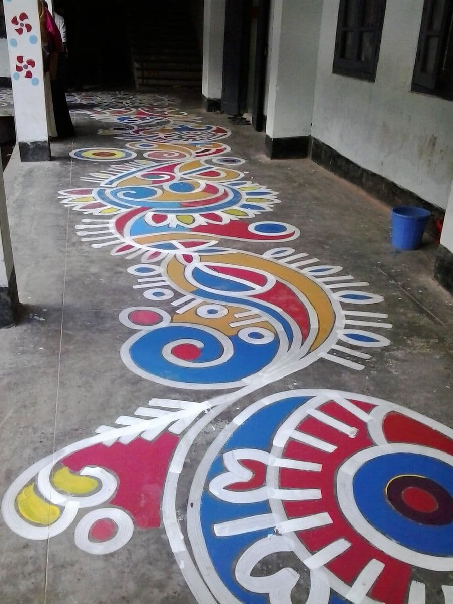 A polychrome alpona running the length of a covered corridor, white outlines filled with red, blue and yellow, with Bengali lettering.