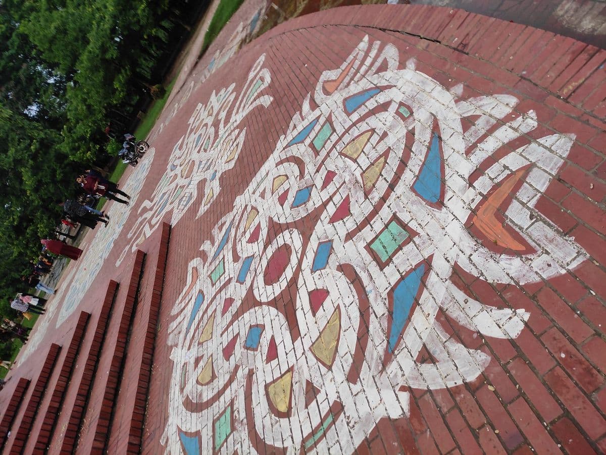 A vast alpona covering the brick paving before the Shaheed Minar at Jahangirnagar University, Bangladesh, with visitors walking across it.