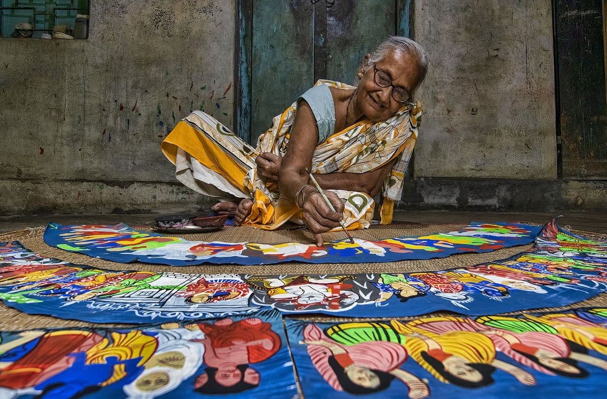 An elderly Krishnanagar painter at work on the floor, filling long curved chalchitra strips with flat colour by hand.