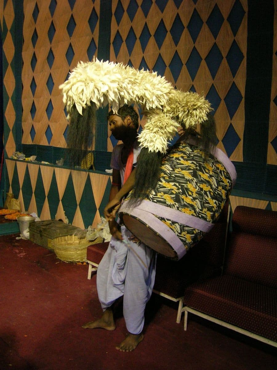 A plumed dhak slung high on the player's chest at a south Kolkata pandal, white feather sprays and long fringes attached.