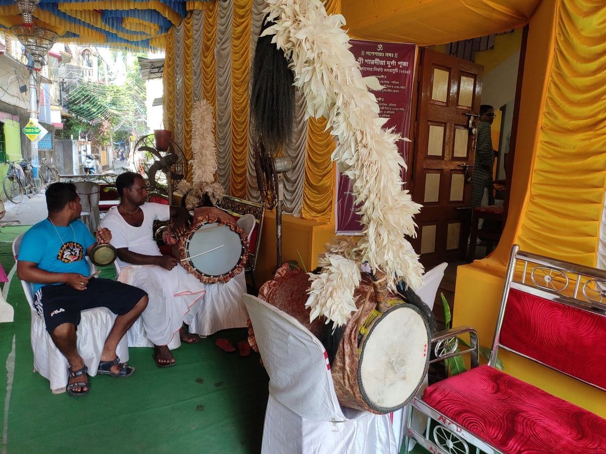 Two musicians in a pandal with a kansar plate and a dhol, two plumed dhaks standing ready beside them.