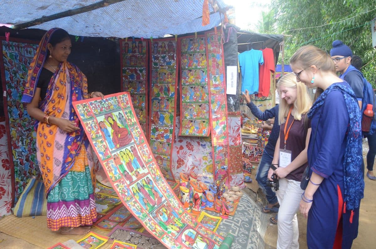 A Naya patua unrolls a long jorano pat for foreign visitors at a village stall hung with painted scrolls.