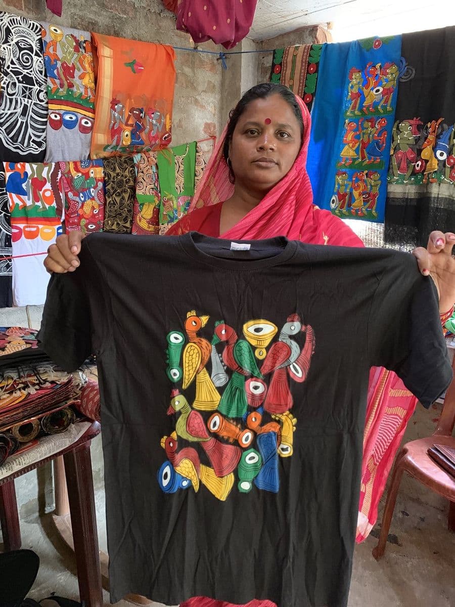 The patua Joba Chitrakar holds up a hand-painted T-shirt in Pingla, with painted saris and stoles hanging behind her.