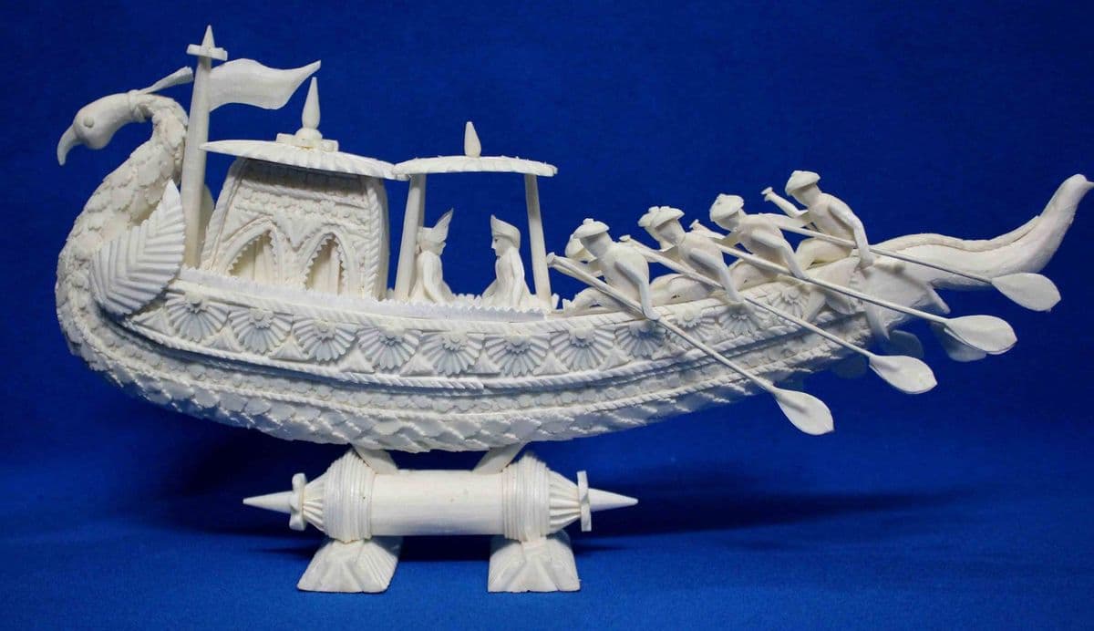 A peacock-prowed boat with rowers, oars and canopy, carved entirely from white shola pith without moulds or templates.