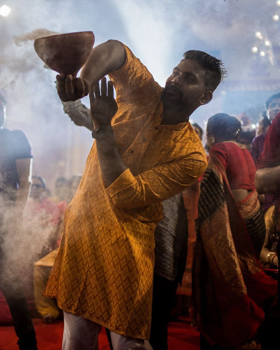 A dancer in a printed kurta swings a burning dhunuchi above his head, the crowd pressed close around him in smoke.